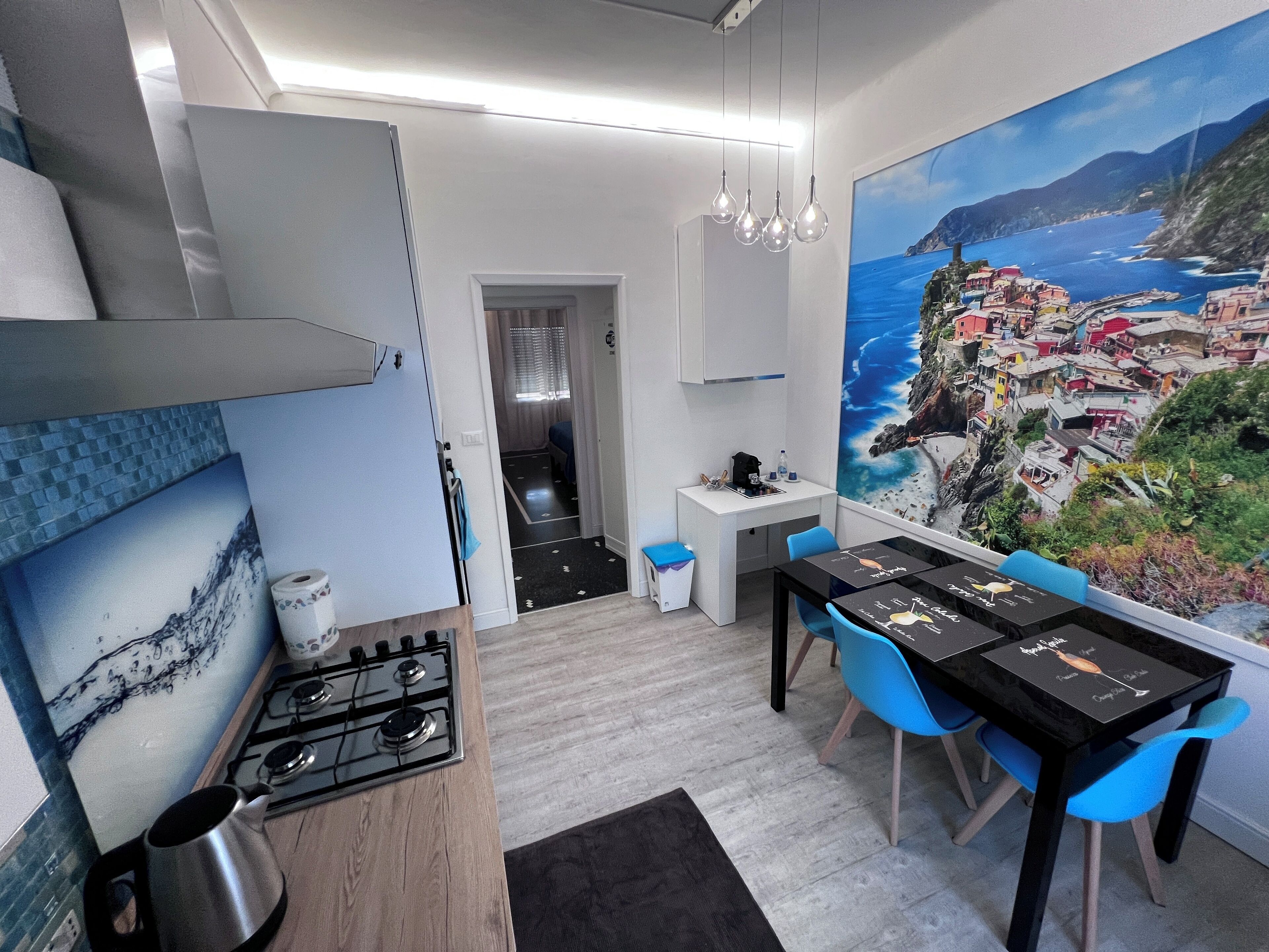 Eine moderne Küche mit Gasherd, Spüle und Essbereich inklusive Tisch und Stühlen. An der Wand hängt ein Gemälde einer Küstenstadt.
