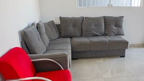 Un sofá seccional gris con varios cojines, una silla roja con estructura metálica y un piso de baldosas.