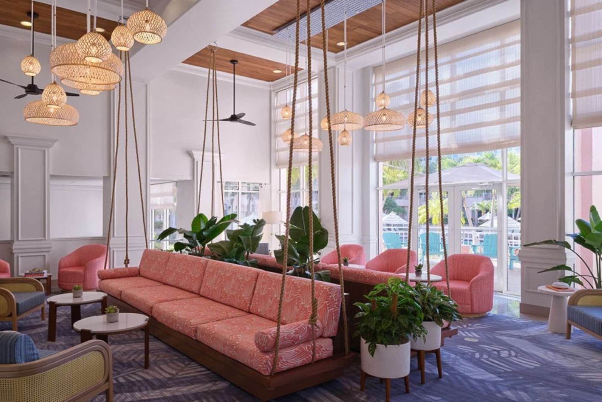 A spacious lounge area with a large sofa, multiple chairs, hanging lights, and large windows offering a view of the outdoors.