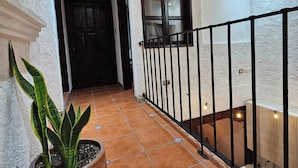 A hallway with terracotta tiles, a potted plant, a window with curtains, and a staircase with a black railing.