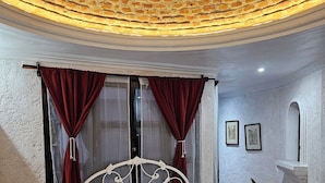 A bedroom with a brick ceiling, a bed with white linens, red curtains, and a bedside table with a lamp.