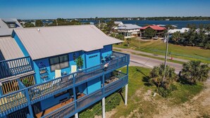 A multi-story building with a blue exterior, a balcony with outdoor seating, and a view of a body of water and distant buildings. - Gulf Shores West Beach Condo/3-Min Walk to Beach/Gulf Views/King Bed (Gulf Shores)