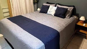 A bed with a blue and white bedspread, a wooden nightstand with a lamp, and a wall with a curtain. - Apartment in João Pessoa (Pedro Gondim João Pessoa)
