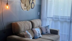 A beige sofa with three cushions, a wall-mounted light fixture, and a window with sheer curtains. - Apartment in João Pessoa (Pedro Gondim João Pessoa)