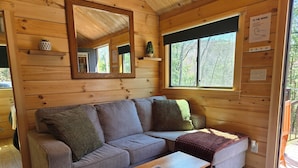 A wooden cabin interior with a sofa, a coffee table, and a window with a view of trees. - Rivertop Retreat, with hot tub!- Luxurious tiny home in the White Mountains! (Bath)