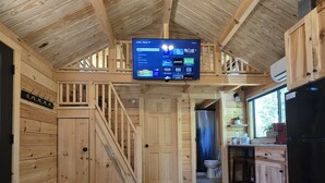 A wooden interior with a TV on the ceiling, a table with remote controls, and a kitchen area. - Rivertop Retreat, with hot tub!- Luxurious tiny home in the White Mountains! (Bath)