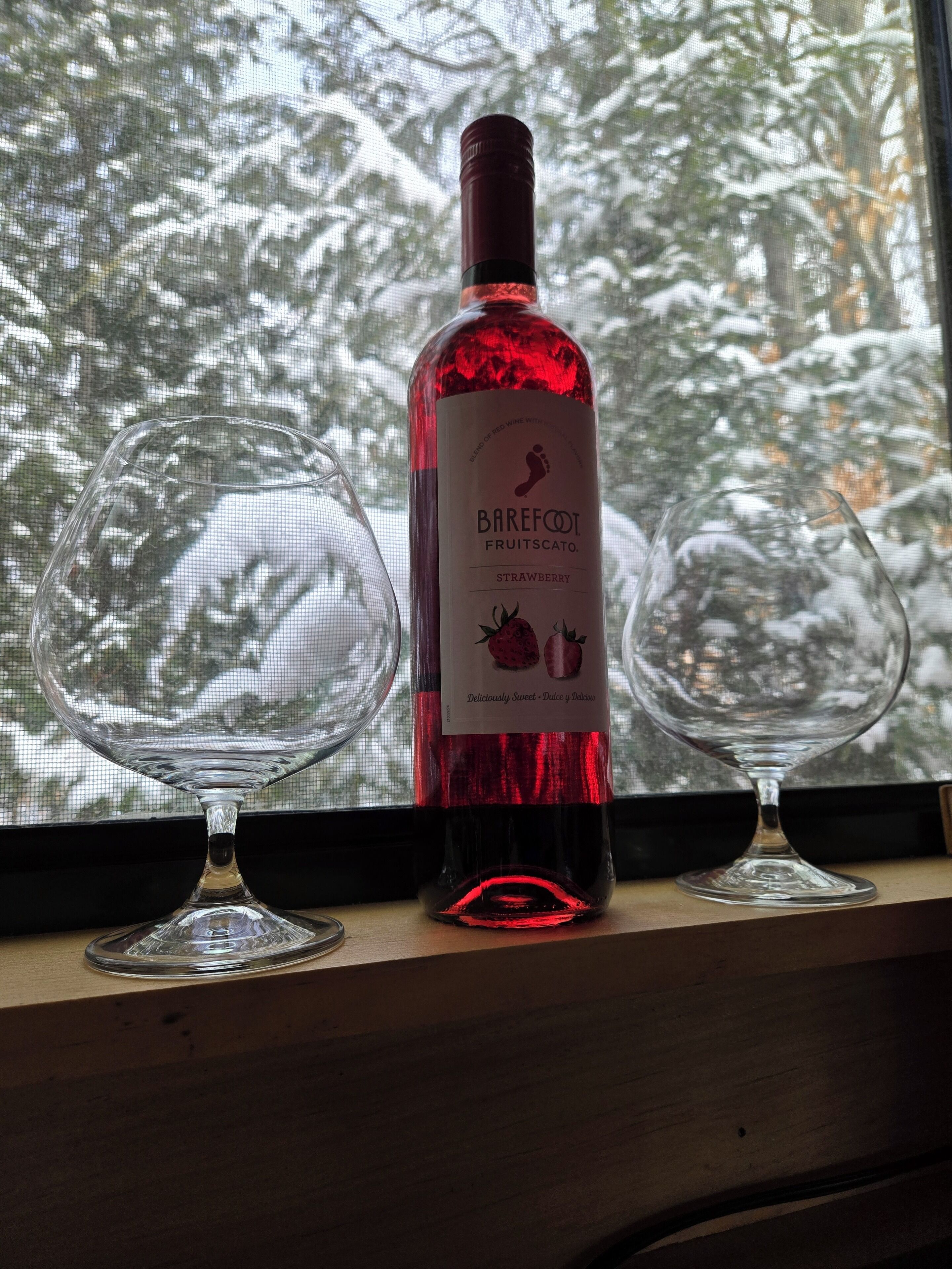 Una botella de vino Barefoot Fruitscato sabor fresa sobre un alféizar con dos copas de vino vacías.