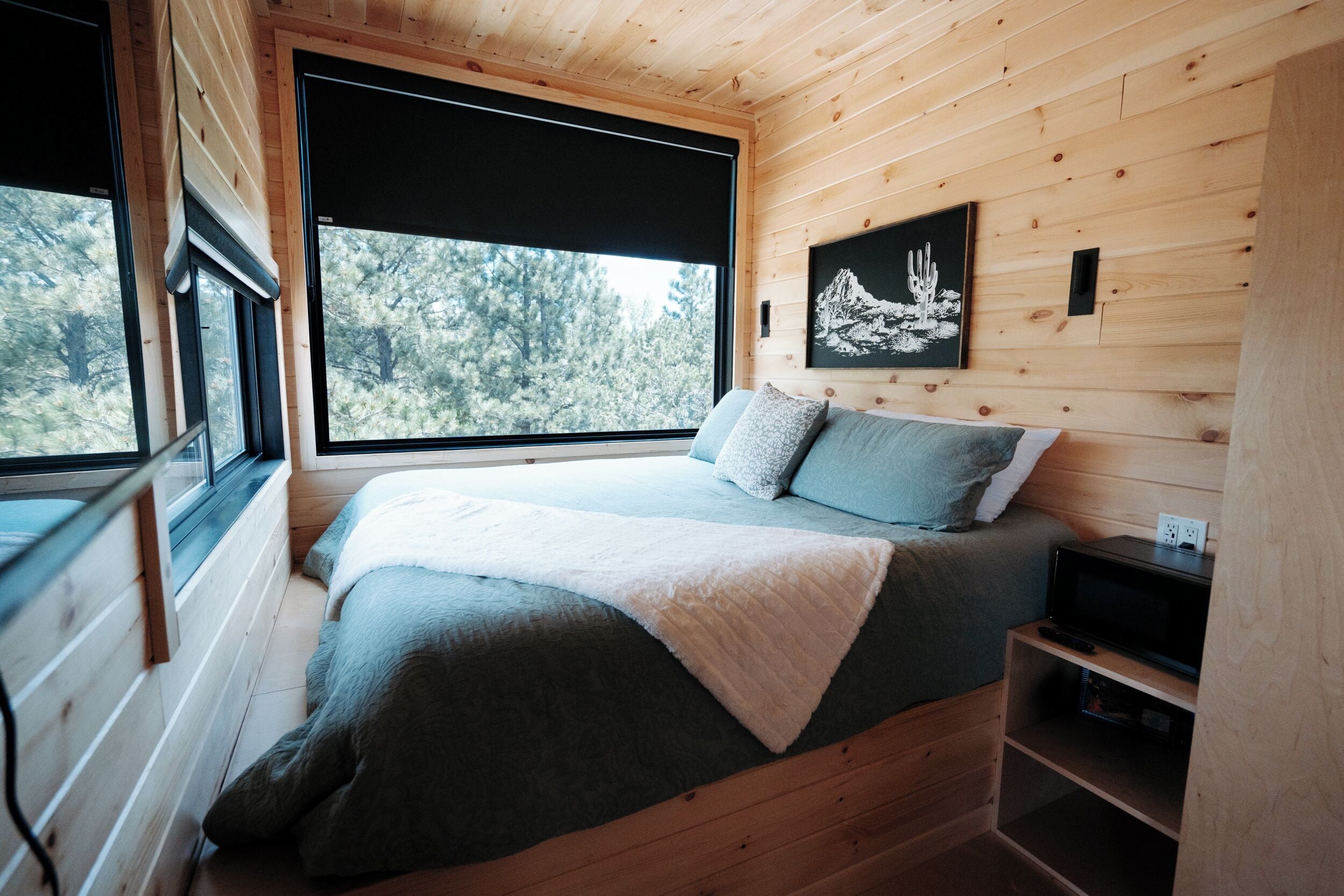 A wooden cabin bedroom with a bed, a window, a painting, and a shelf.