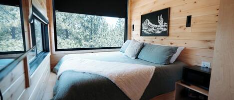 A wooden cabin bedroom with a bed, a window, a painting, and a shelf.