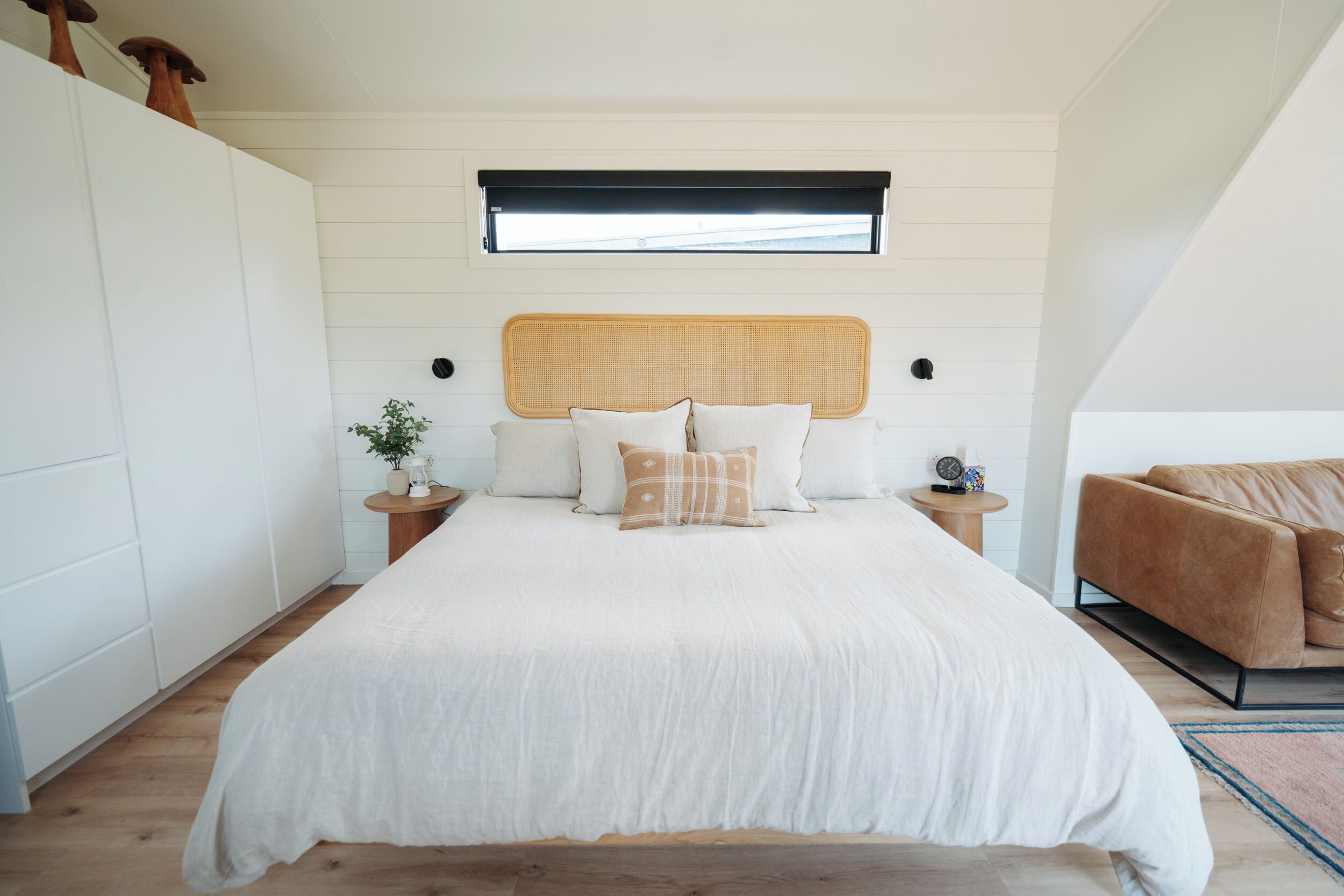 A bedroom with a large bed, a headboard, bedside tables, a potted plant, and a brown sofa.