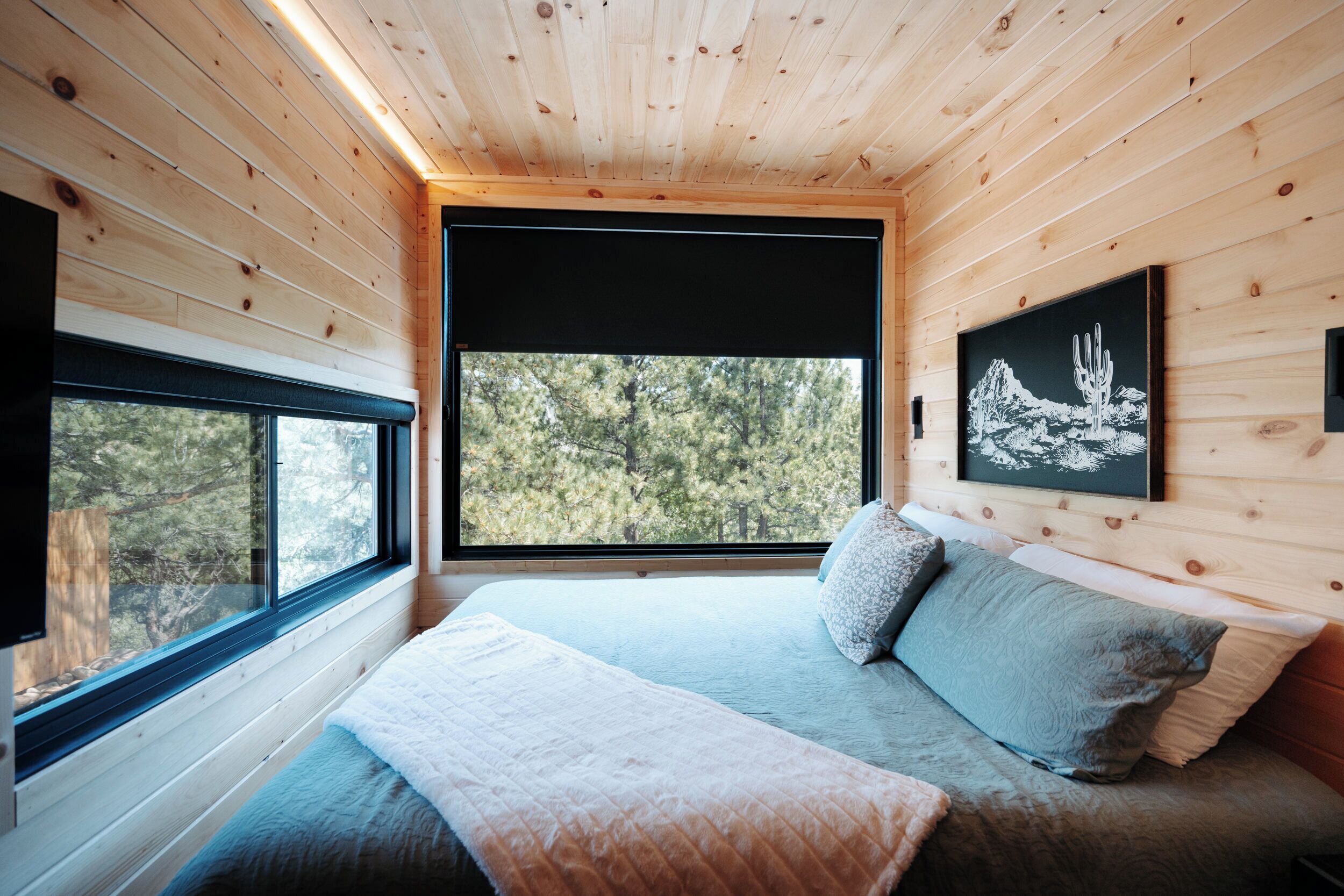 A wooden cabin interior with a bed, large windows, and a framed picture on the wall.