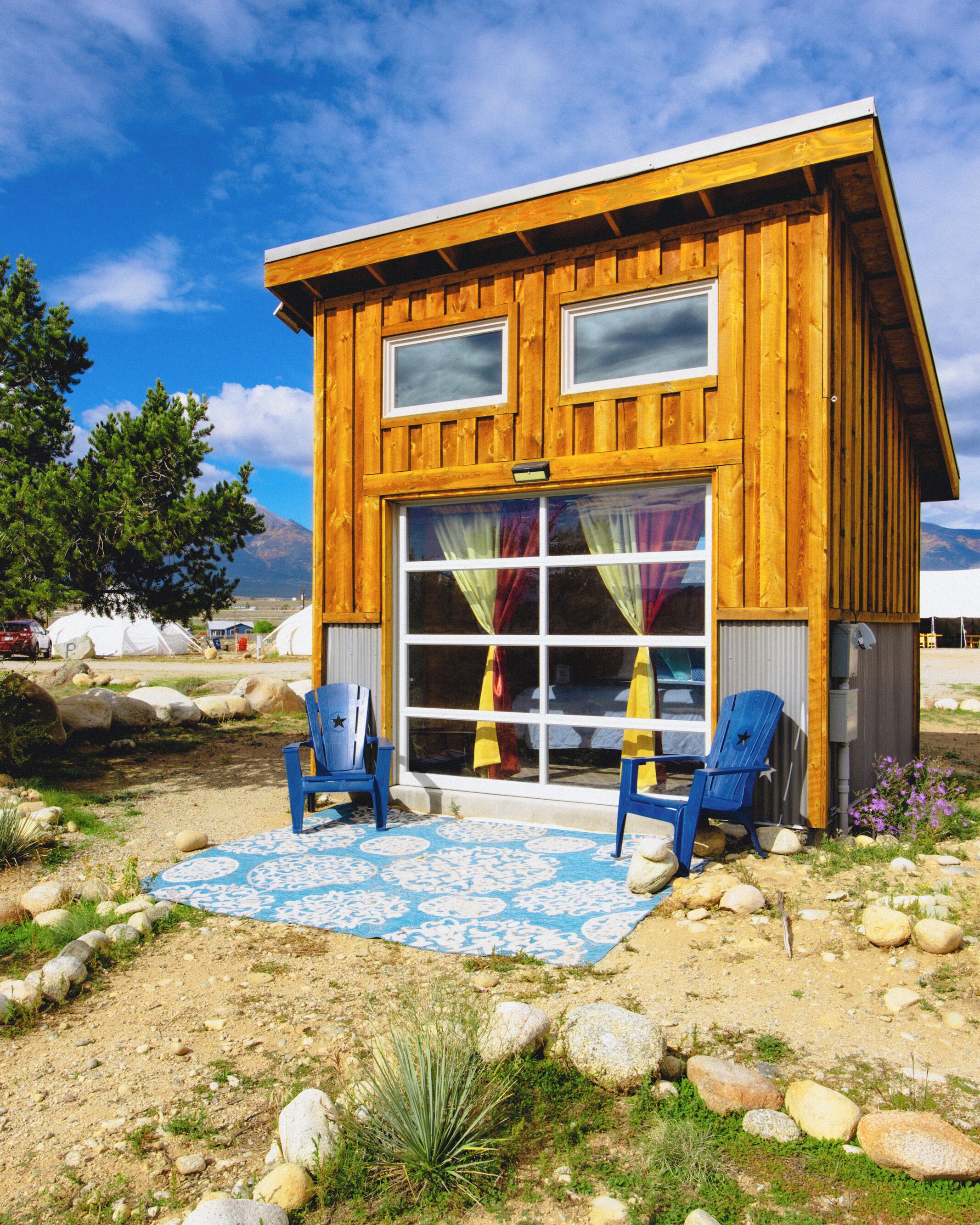 Une cabane en bois avec deux fenêtres, une grande porte coulissante et deux chaises bleues à l’extérieur.