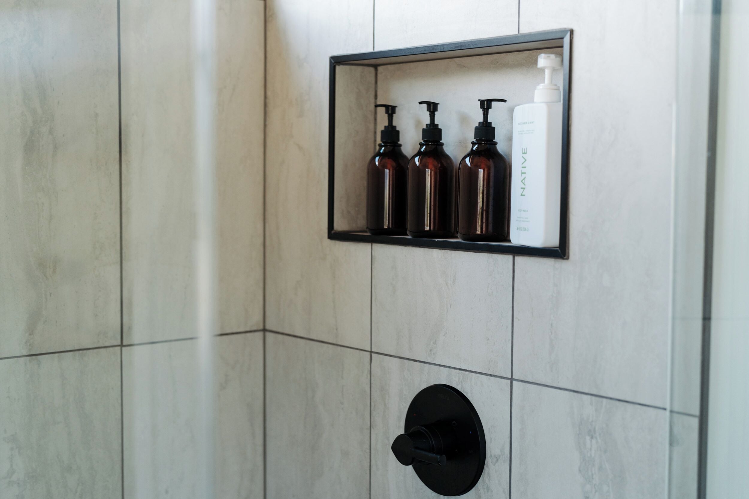 A bathroom with a tiled wall, a black sink, and a shelf with three bottles.