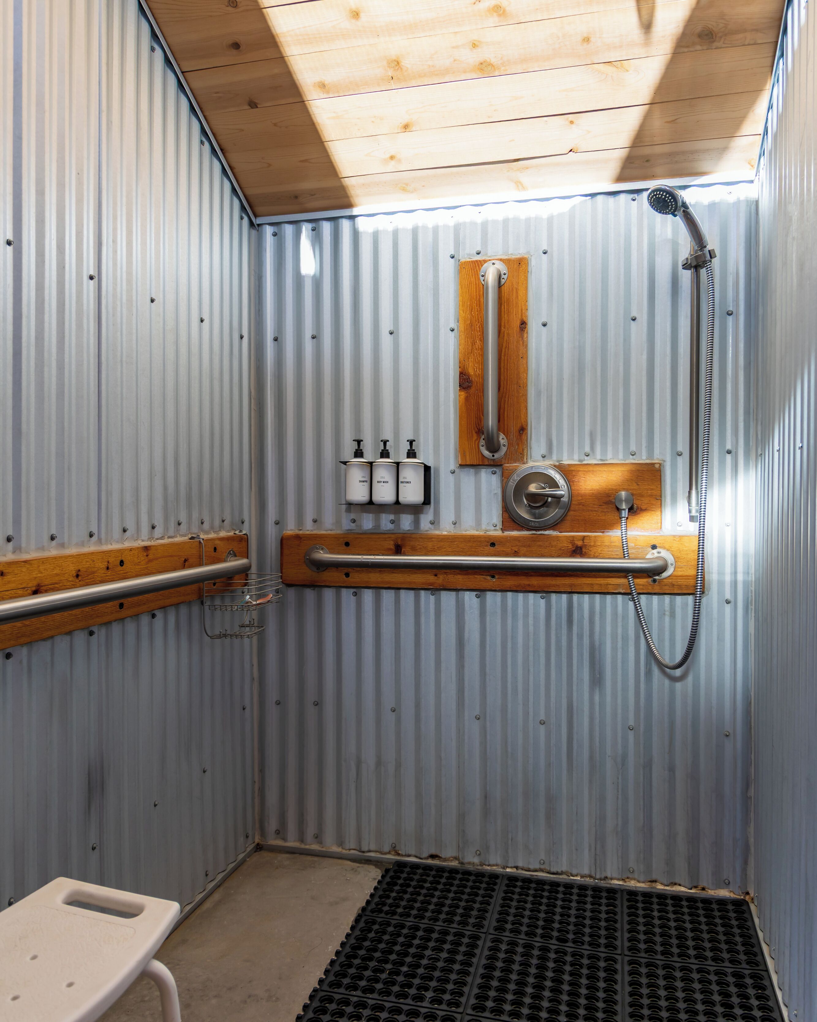 Un baño pequeño con paredes de metal, techo de madera, ducha, inodoro y un estante con artículos de aseo.