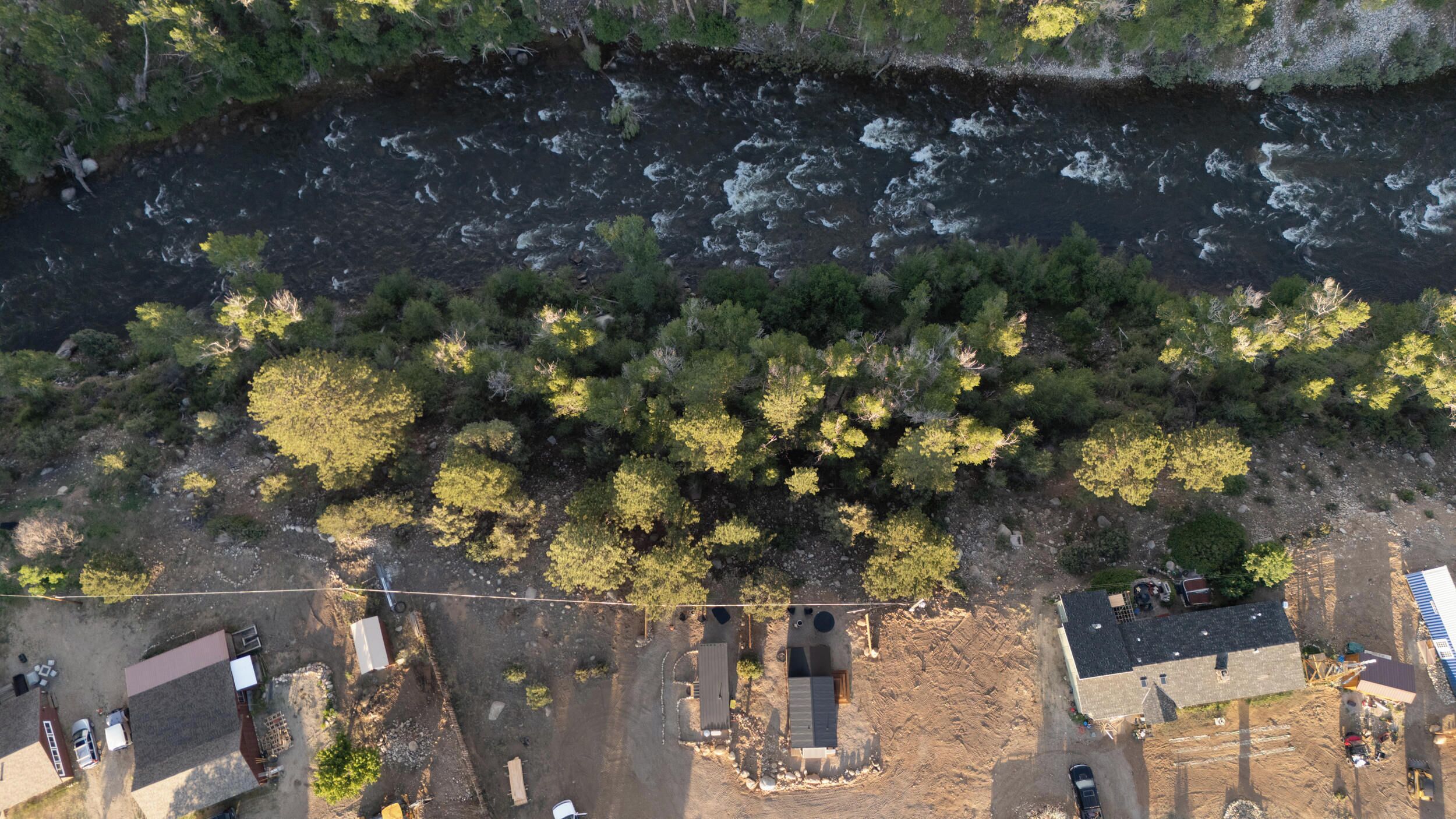 An aerial view of a riverside property with a dirt road, a small building, and a large tree-covered area.