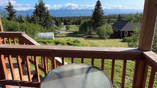 A wooden deck with a round table and chairs, overlooking a residential area with houses and a mountain range.