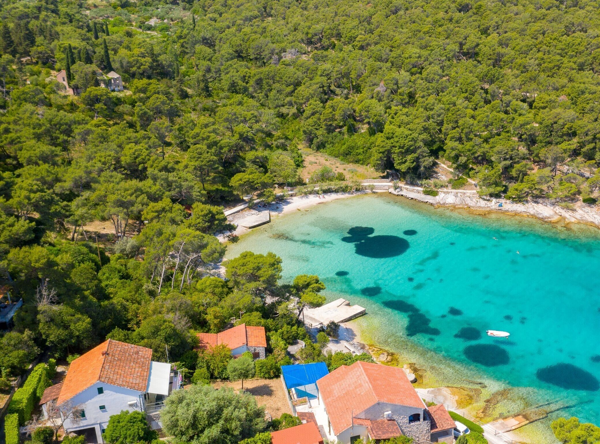Une vue aérienne d’une zone côtière aux eaux turquoise et limpides, bordée d’une forêt verdoyante et dense, et où l’on aperçoit plusieurs bâtiments aux toits recouverts de tuiles rouges.