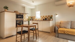 Una cuina compacta amb armaris blancs, taula de dinar de fusta i un sofà a la zona de sala d'estar.