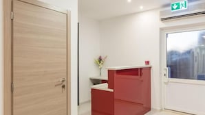 Un área de recepción con una puerta de madera, un mostrador rojo, un florero con flores y una puerta de vidrio con letrero de salida verde.