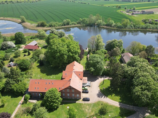 Oversigtsbillede af en stor ejendom med en markant bygning med rødt tag, omgivet af frodig vegetation og en sø.