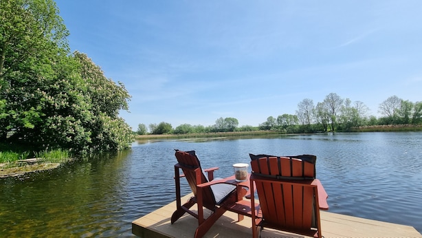 Eine Holzstele mit zwei Stühlen und einem Tisch am Ufer eines ruhigen Flusses, umgeben von üppiger Vegetation und Bäumen.