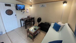 A compact living space with a dining area, a TV mounted on the wall, a fan, and a couch with a pillow. - Cozy 1-bedroom apartment with WiFi, Pool near the Airport & Store Bay Beach (Crown Point)