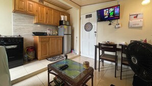 A compact kitchen with wooden cabinets, a refrigerator, a stove, and a dining area with a table and chairs. A television is mounted on the wall. - Cozy 1-bedroom apartment with WiFi, Pool near the Airport & Store Bay Beach (Crown Point)