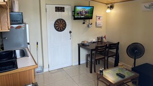 A compact living space with a kitchen area, a dining table with chairs, a dartboard, a television mounted on the wall, and a fan. - Cozy 1-bedroom apartment with WiFi, Pool near the Airport & Store Bay Beach (Crown Point)