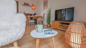 A cozy living room with a fluffy white chair, a small round table with a magazine, a television, and a wooden cabinet. - Refugium Möhneseeblick. our cozy vacation apartment.central and yet quiet. (Möhnesee)