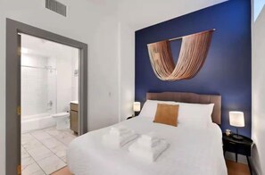 A hotel room with a bed, bedside lamps, a hanging textile decoration, a shower area, and a visible bathroom.