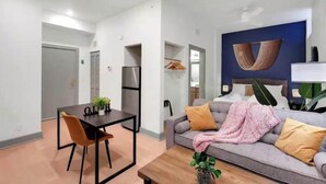 Una habitación de hotel moderna con comedor, sofá con cojines, una planta grande y una obra de arte colgada en la pared.