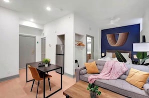 Una habitación de hotel moderna con comedor, sofá con cojines, una planta grande y una obra de arte colgada en la pared.