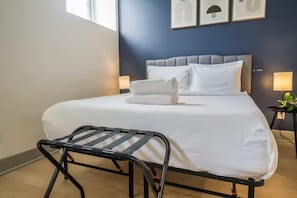 Un lit bien fait, avec du linge de lit blanc, une tĂȘte de lit rembourrĂ©e de couleur grise et une chaise en mĂ©tal noir placĂ©e devant.