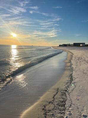 Une plage avec un ciel dégagé, le soleil se couchant ou se levant, et des bâtiments en arrière-plan.