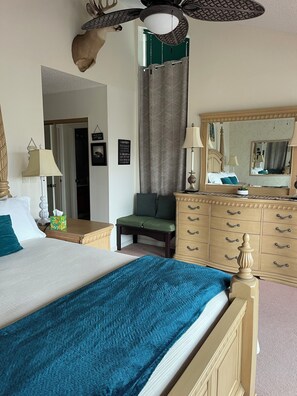 Una habitación de hotel con una cama, un aparador, una silla, un espejo y un ventilador de techo.