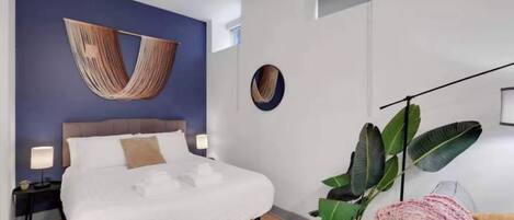 A modern bedroom with a bed, bedside lamps, a large plant, and a unique wall hanging.