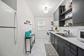 A kitchen with white tiled walls, a black countertop, and a window with blinds.