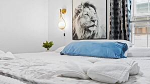 Un letto rifatto con una coperta bianca e blu, una cornice con un'immagine di un leone, un lampadario a parete e una finestra con tende.
