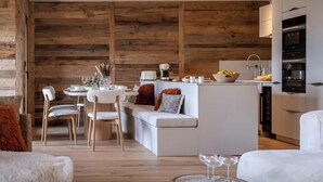 A modern kitchen with a wooden wall, a dining area with chairs and a table, and a sofa with a fluffy pillow.