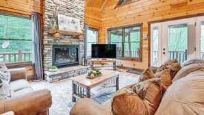 A cozy cabin interior with a stone fireplace, wooden walls, and large windows offering a view of the forest.