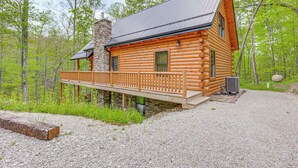 A log cabin with a wooden deck, stone chimney, and a gravel driveway.