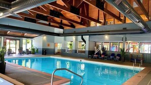 An indoor swimming pool with a wooden ceiling, clear glass walls, and a seating area with chairs and a table.