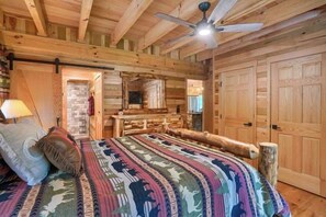 A log cabin bedroom with a bed, wooden walls, a ceiling fan, and a large mirror.