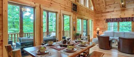 A wooden cabin interior with a dining table set for four, a sofa, and a large window overlooking greenery.