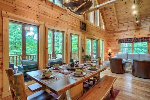 Interior de una cabaña de madera con una mesa de comedor lista para cuatro personas, un sofá y un ventanal con vistas a la vegetación.