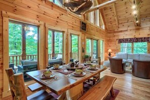 A wooden cabin interior with a dining table set for four, a sofa, and a large window overlooking greenery. - Bear Pines charming 3 bdrm cabin, Hot Tub, WiFi, in scenic South Bloomingville (South Bloomingville)