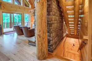 A log cabin interior with a stone fireplace, wooden staircase, and a view of the outdoors.