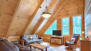 A cozy wooden cabin with a loft bedroom, a living room with a sofa and armchair, a television, and a dining area.