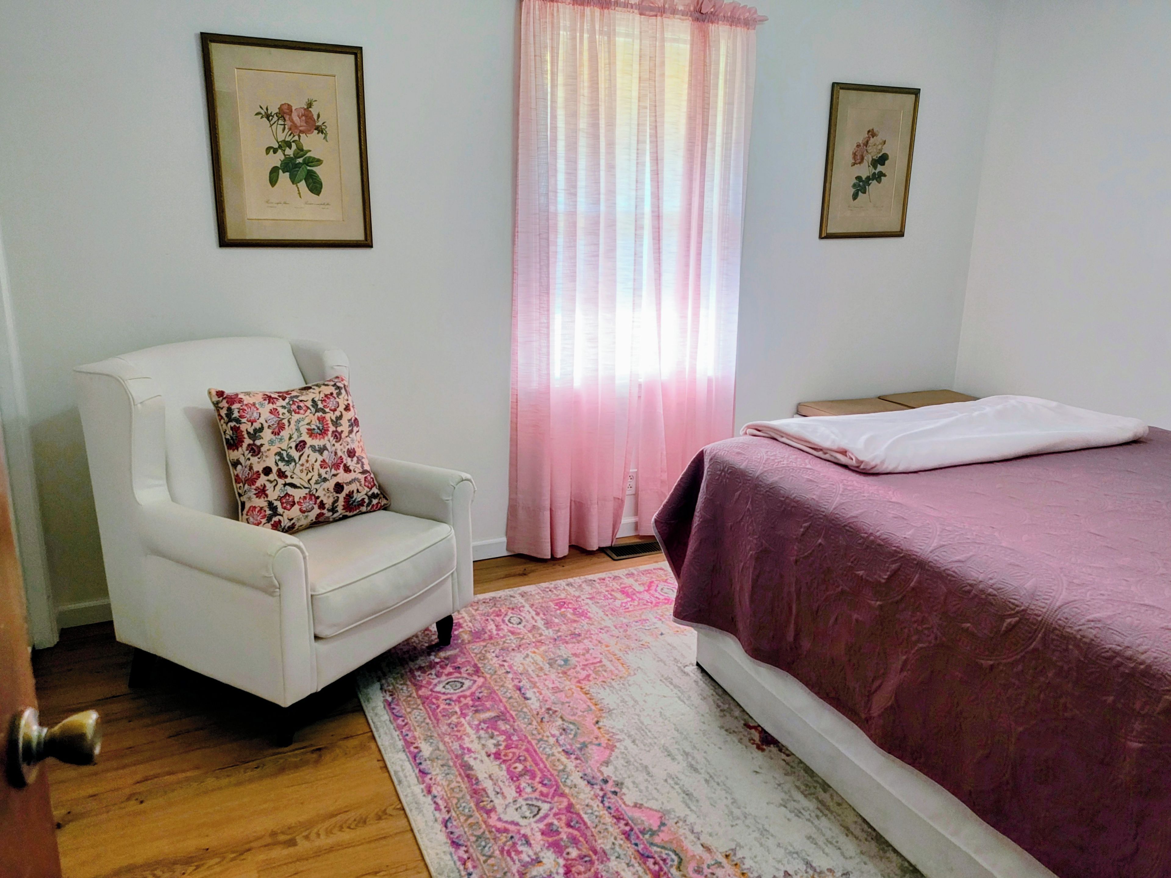 Un dormitorio con un sillón blanco, una cama con colcha rosa, un tapete estampado y dos láminas botánicas enmarcadas en las paredes.