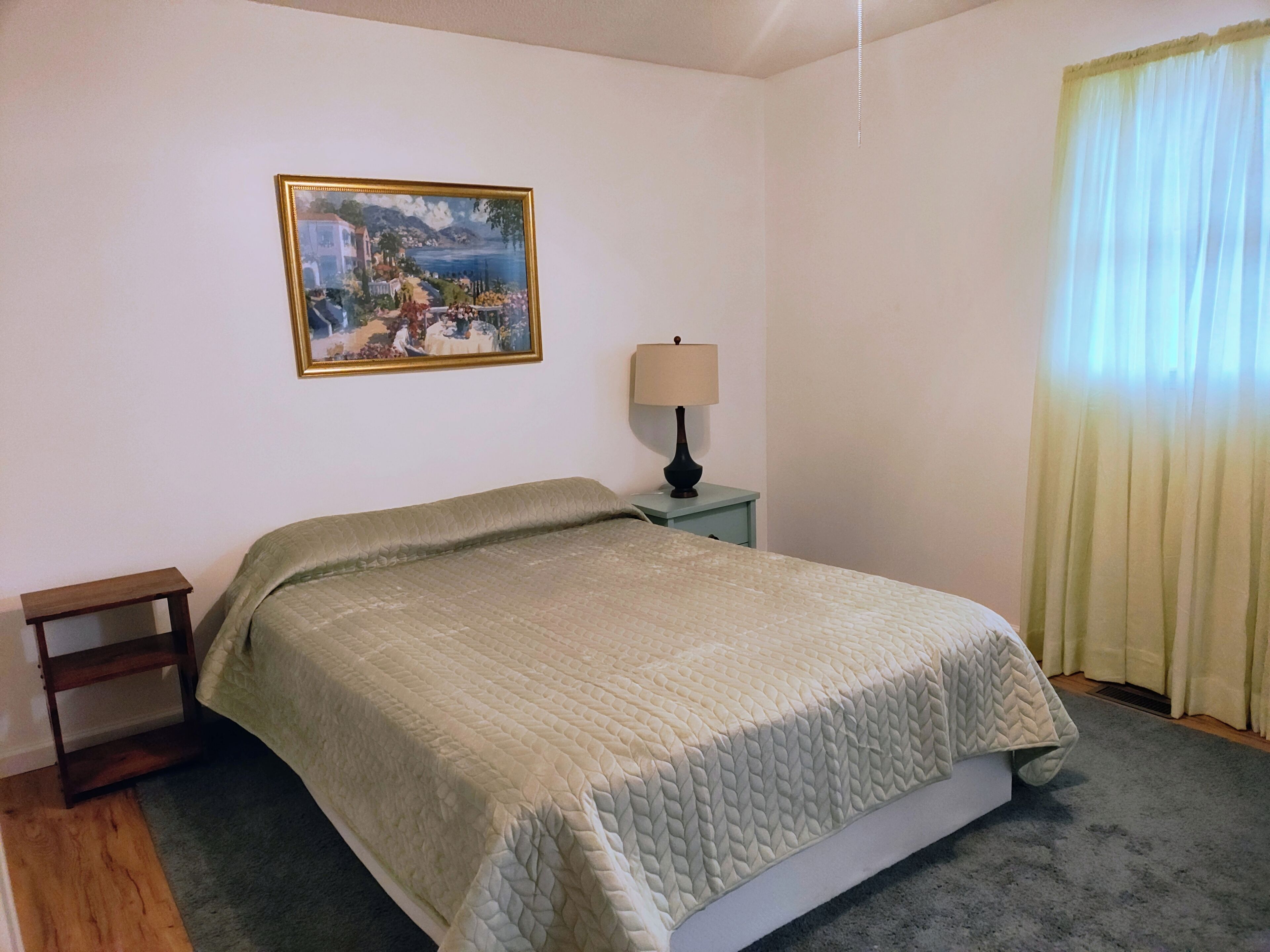 Un dormitorio con cama, mesita de noche, lámpara, un cuadro en la pared y una ventana con cortinas.